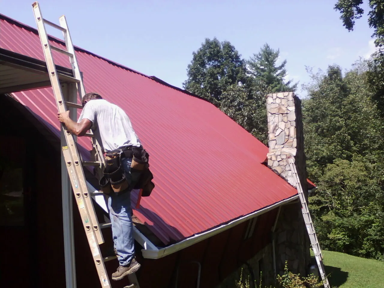 Roofer on a ladder beside a red metal roof and stone chimney — Soffit & Fascia Repair by Roofing Brothers in Peralta